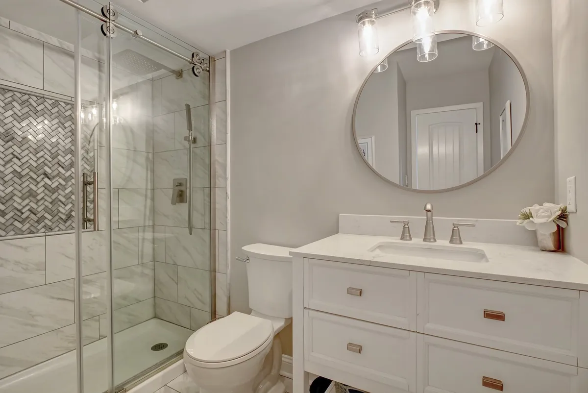 A modern bathroom with a glass shower enclosure, a stylish vanity with sink, a round mirror, and a decorative vase with flowers. The space is refreshing, well-lit, and designed for both morning routines and relaxing baths.