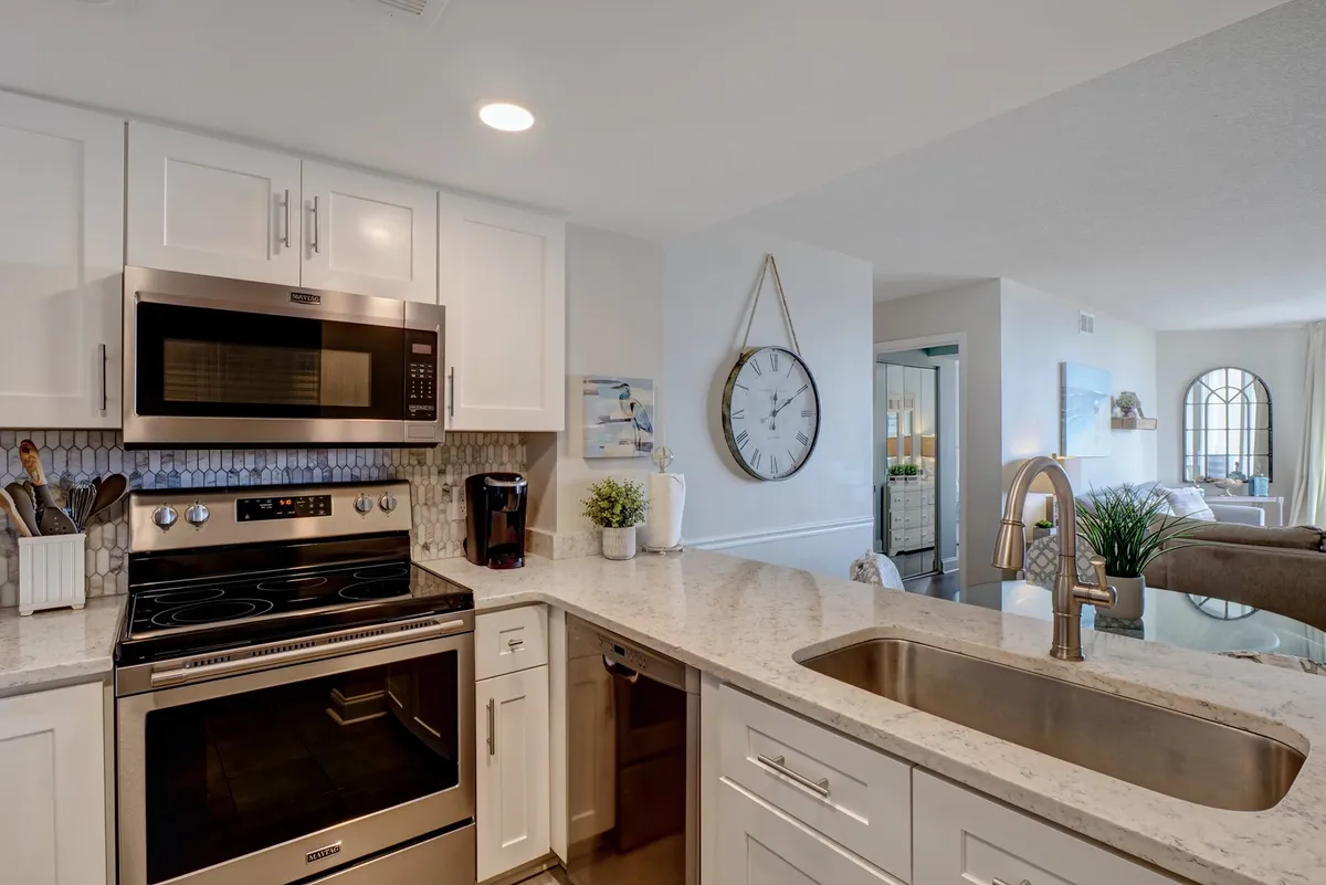A contemporary kitchen with stainless steel appliances, white cabinetry, granite countertops, a decorative wall clock, and a view into the adjacent living area. The design is clean, modern, and inviting—ideal for both everyday meals and entertaining guests.