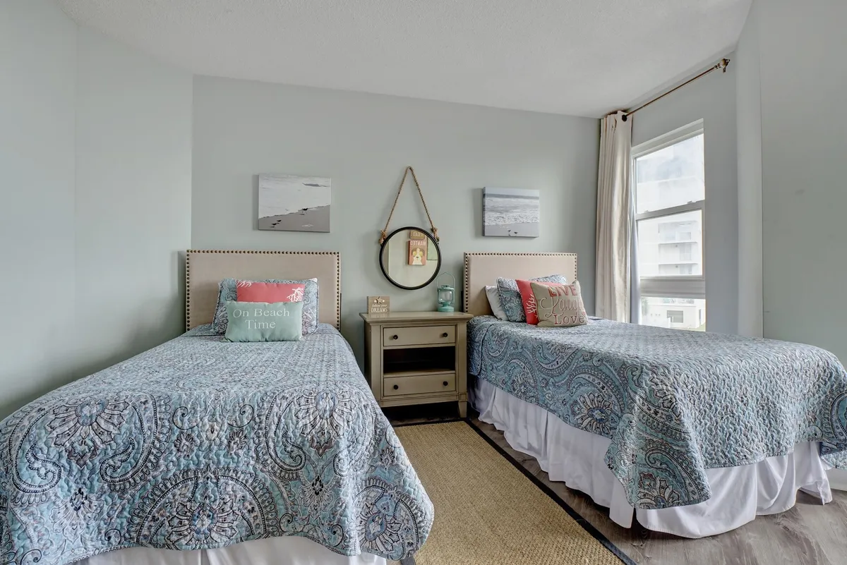 A cheerful bedroom featuring two twin beds adorned with decorative quilts and beach-themed pillows, wall art depicting beach scenes, a round hanging mirror, and a bedside table with a lamp. Sunlight streams into the room, enhancing its inviting, family-friendly appeal.