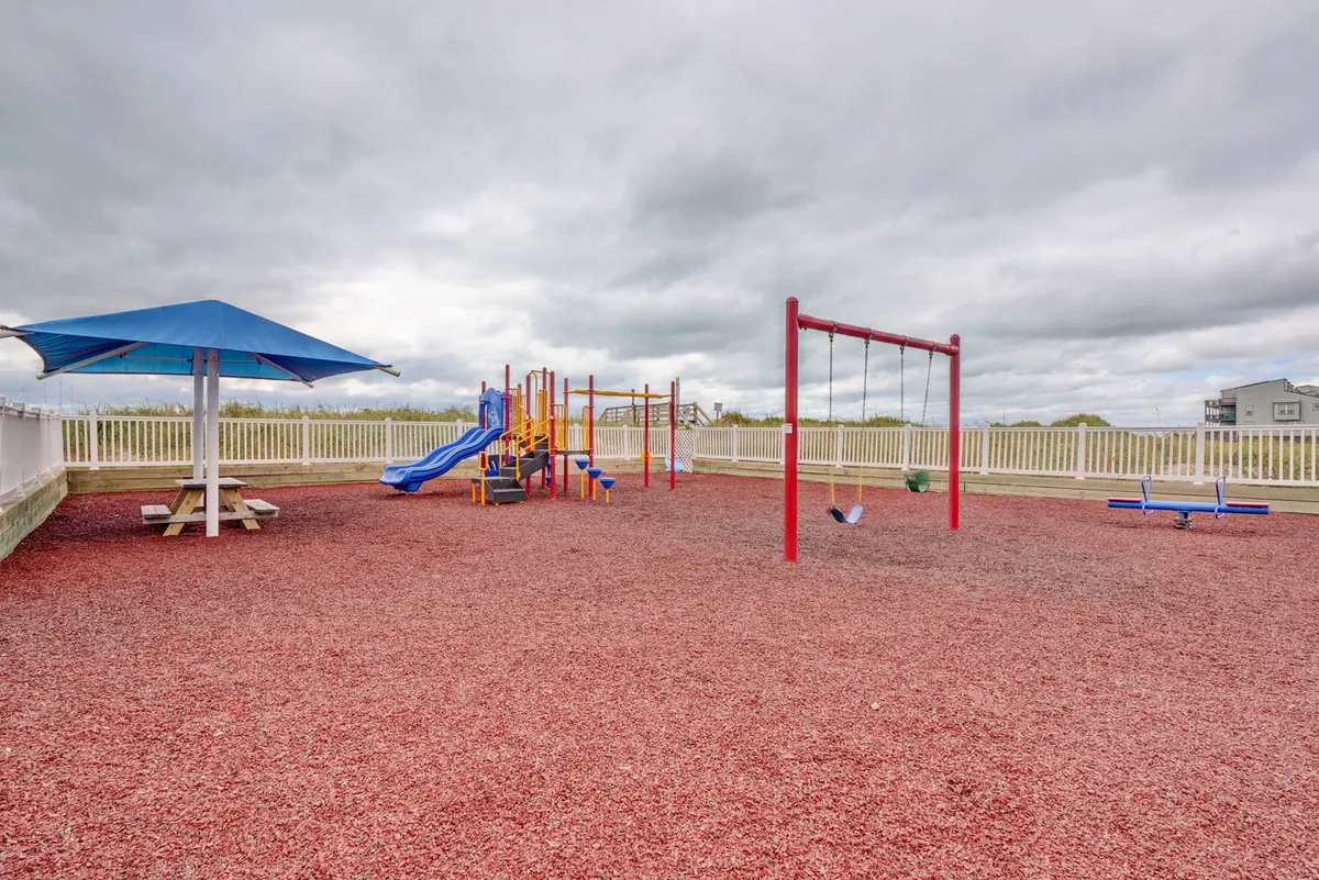 A colorful playground with swings and a picnic table under an umbrella, set in a safe, cheerful outdoor environment. The scene is lively and communal, ideal for families and young guests.