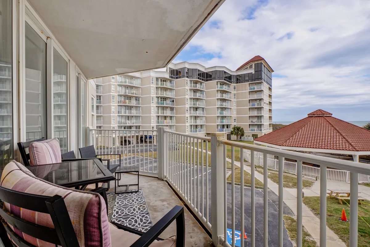 This image presents an inviting balcony with outdoor seating and a sweeping ocean view. Surrounding buildings are visible in the distance, providing context to the resort setting. The mood is tranquil, suggesting moments of solitude or shared experiences watching sunrises and sunsets.