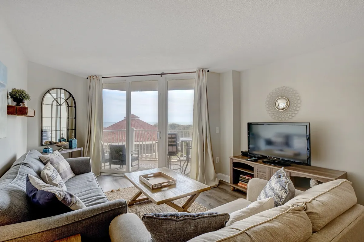 A cozy living room featuring large windows that fill the space with natural light, a comfortable seating arrangement, a stylish coffee table with decor, and views out to a balcony or beyond. The atmosphere is both relaxing and inviting, ideal for unwinding or gathering with family and friends.