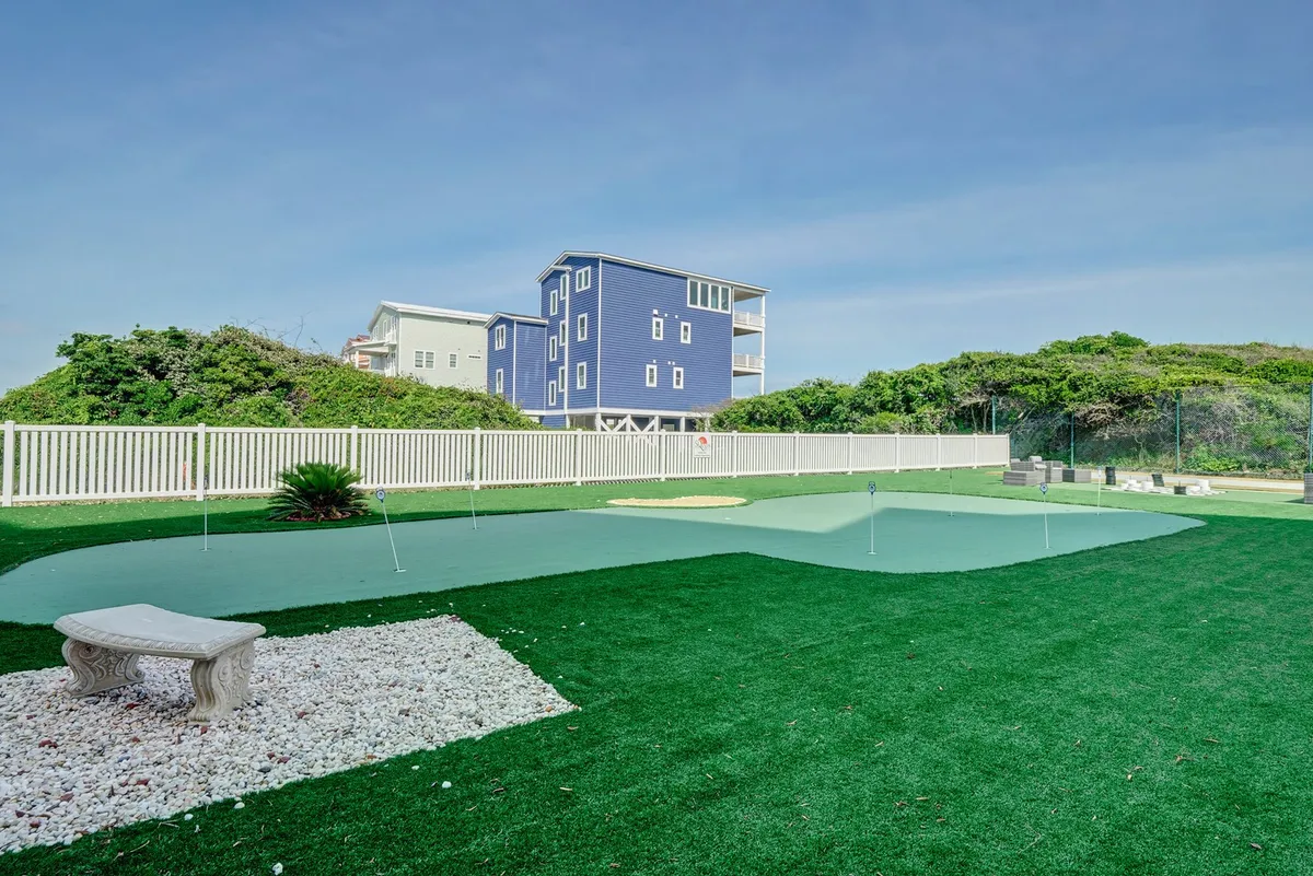 A lush exterior putting green adjacent to a blue house, bordered by landscaping and featuring a stone bench for relaxing between games. The scene is bathed in sunlight, conveying a leisurely atmosphere ideal for group enjoyment and friendly competition.